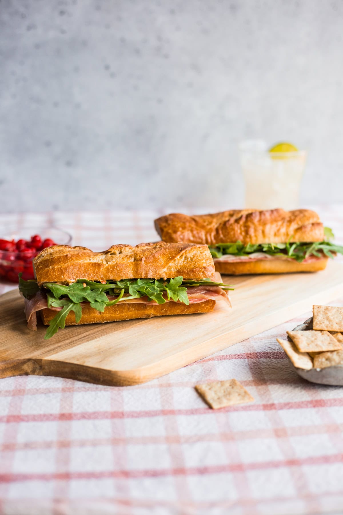 Two baguette sandwiches with arugula, sliced meats, and cheese on a wooden cutting board with crackers in the foreground