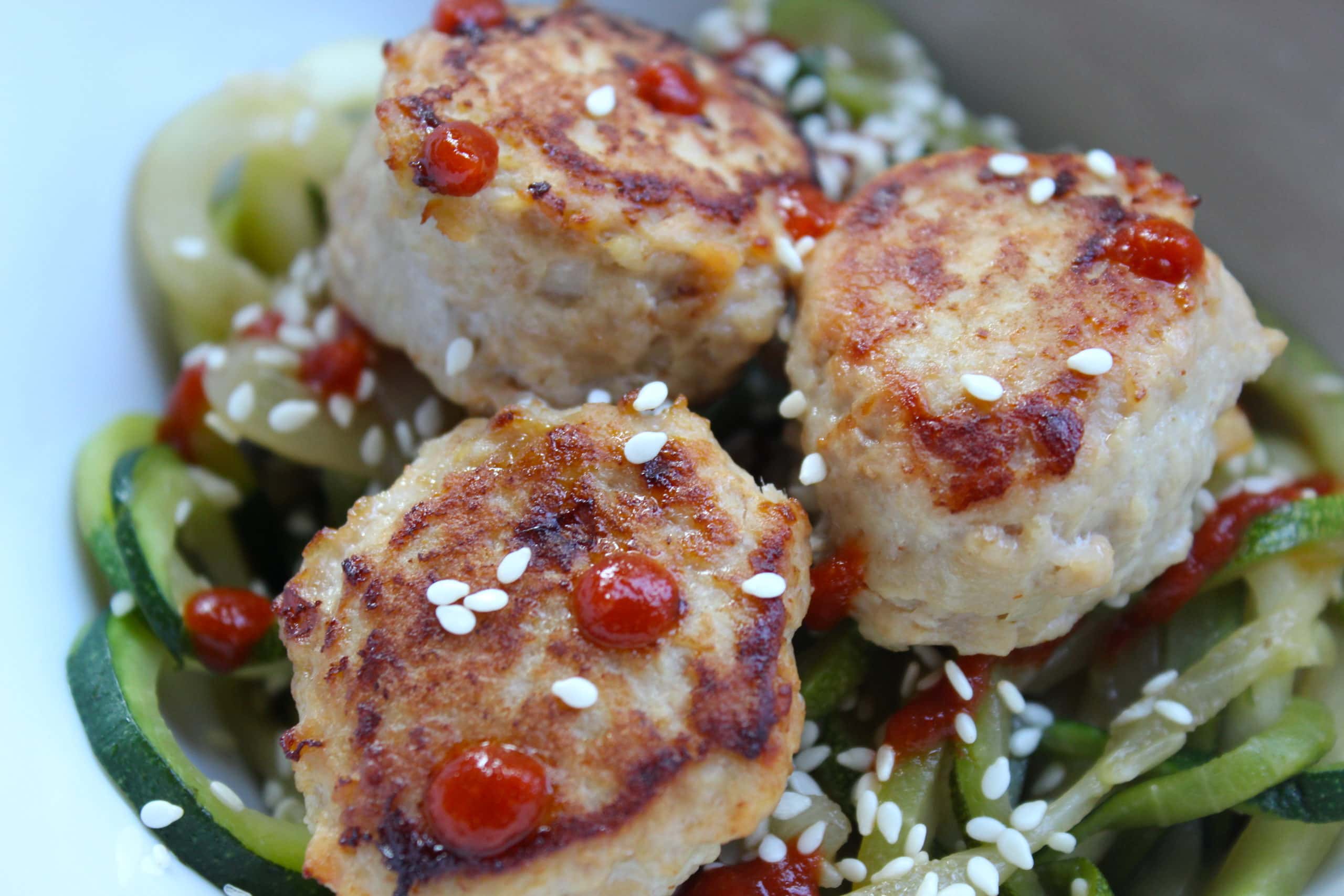 Chicken meatballs topped with sriracha and sesame seeds on top of a bowl of zoodles