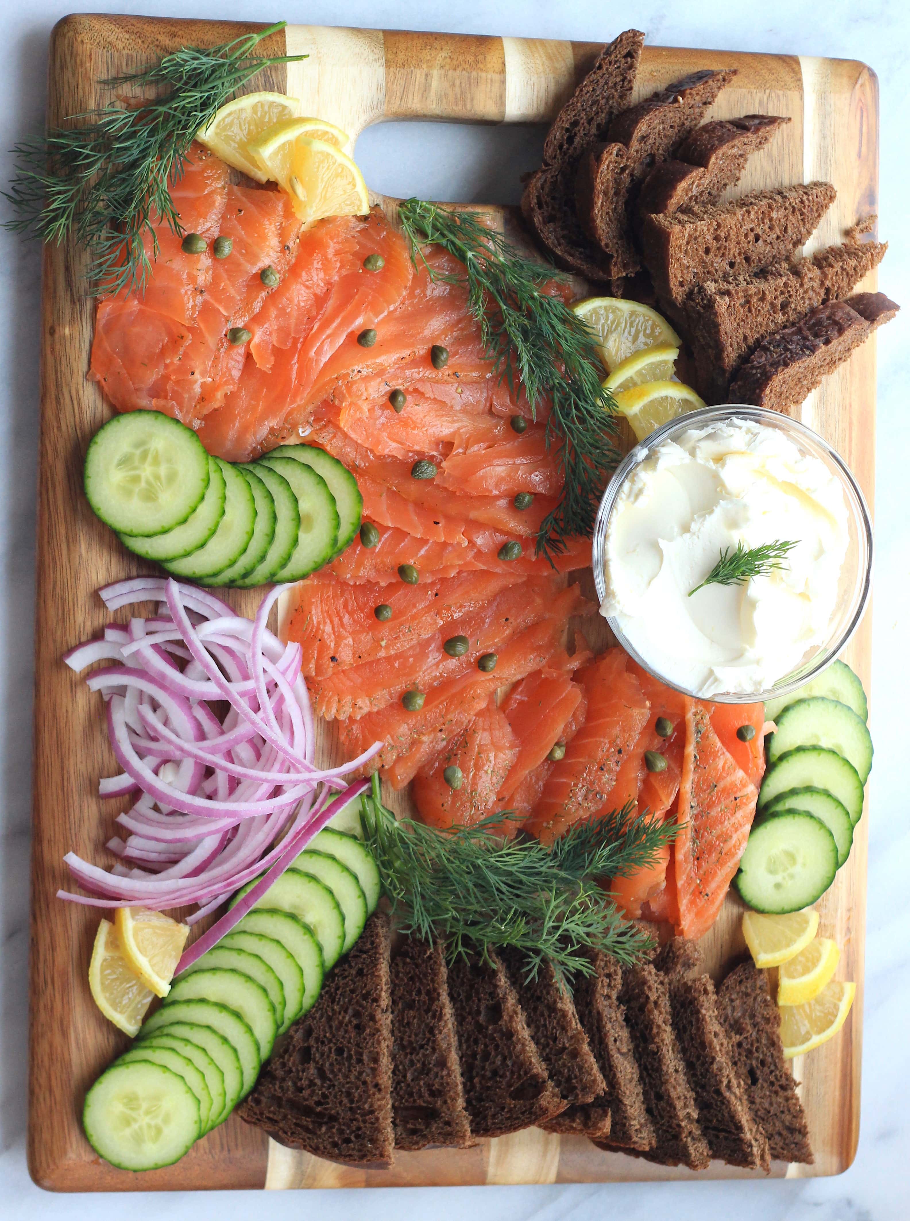 A wooden board topped with lox, capers, red onion, cucumbers, lemon wedges, fresh dill, sliced pumpernickel, and a bowl of cream cheese