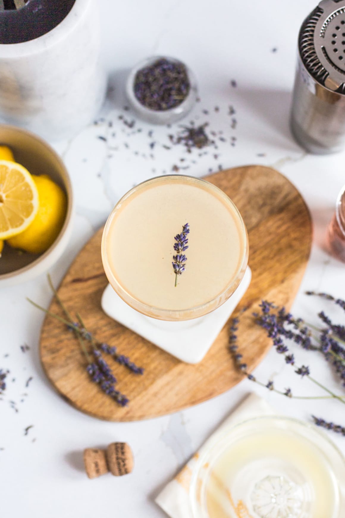A cocktail with a lavender spring in it surrounded by lemons and dried lavender flowers on a wooden cutting board