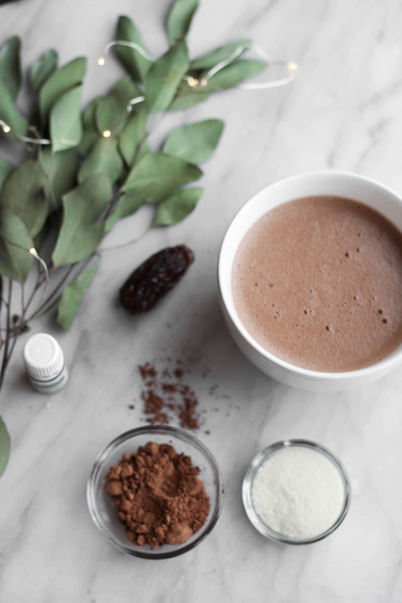 A mug of cocoa near dates, peppermint oil, bowls of cacao, and dried eucalyptus