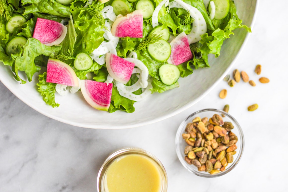 A bowl of greens topped with pink watermelon radish slices and fennel, surrounded by a bowl of pistachios and a jar of yellow vinaigrette