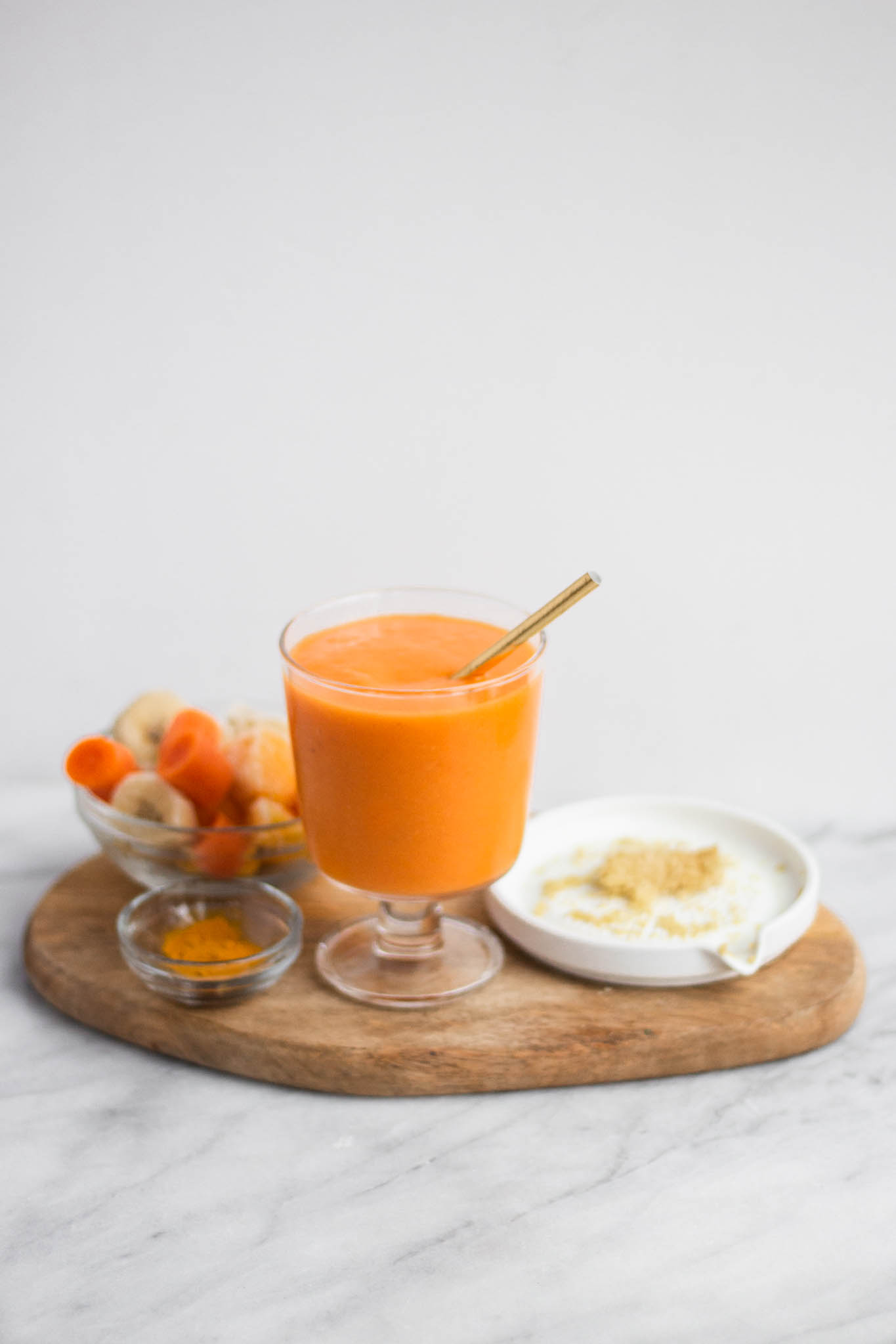 An orange smoothie in a stemmed glass surrounded by grated ginger and frozen mango and carrots