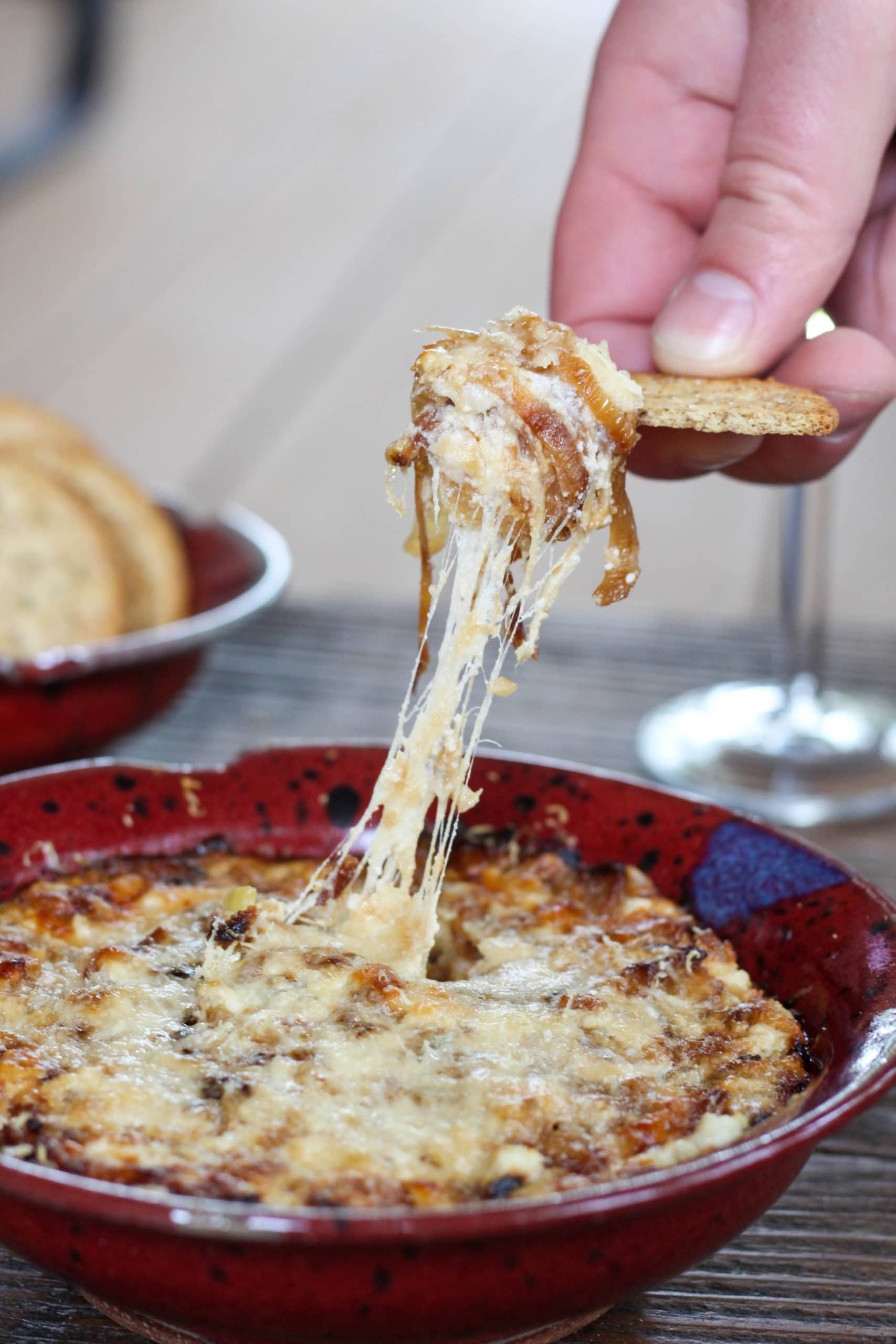 A hand pulling a cracker from a bowl of cheesy caramelized fennel and onion dip