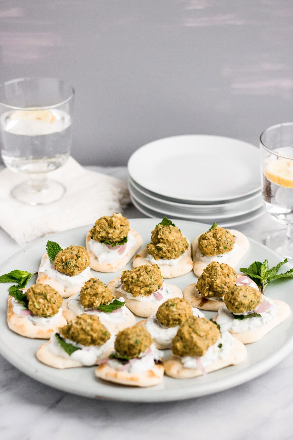 A platter of mini pita bites topped with tzatziki, a mini falafel, fresh mint, and pickled red onion in front of a stack of white plates