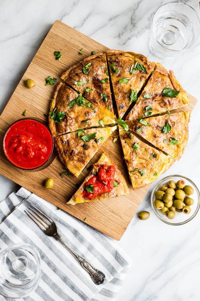 A sliced Spanish Tortilla on a wooden cutting board with parsley and roasted red pepper sauce