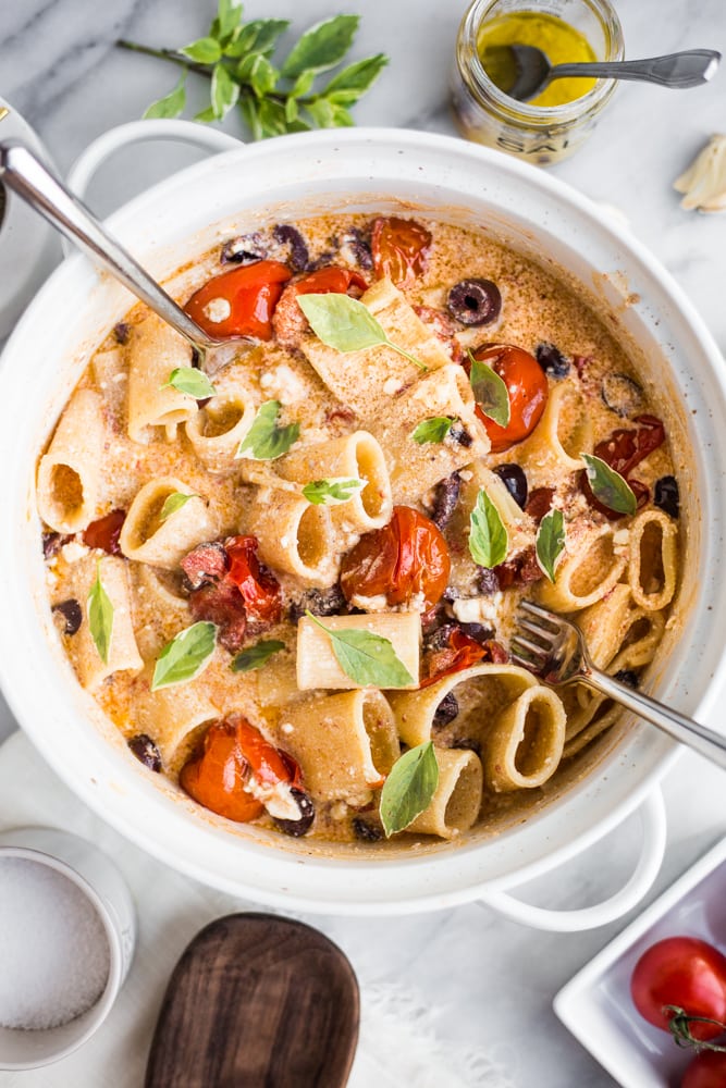 A large bowl with pasta, tomatoes, olives, and basil with two forks in it