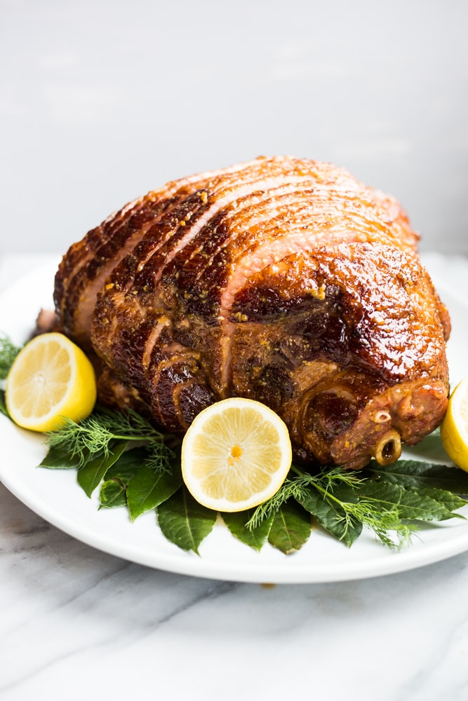 A maple and dijon glazed ham on a platter surrounded by lemon and green herbs