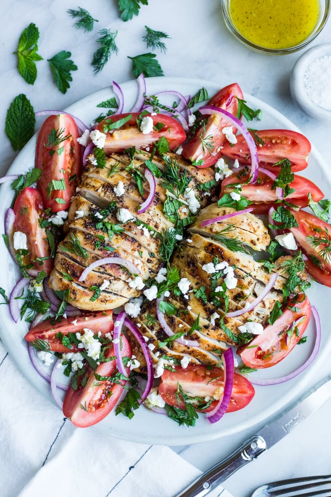 A platter of grilled chicken surrounded bu sliced tomatoes, fresh herbs, red onion, and feta cheese