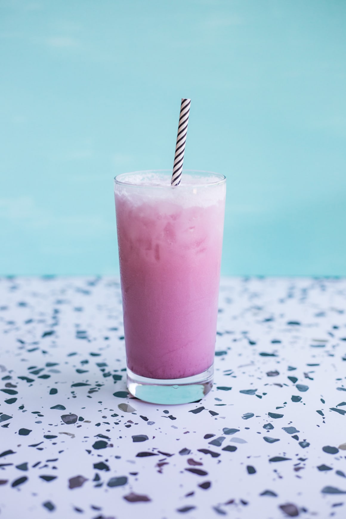 A tall glass filled with a creamy pink beverage with a striped straw, set on a terrazzo countertop