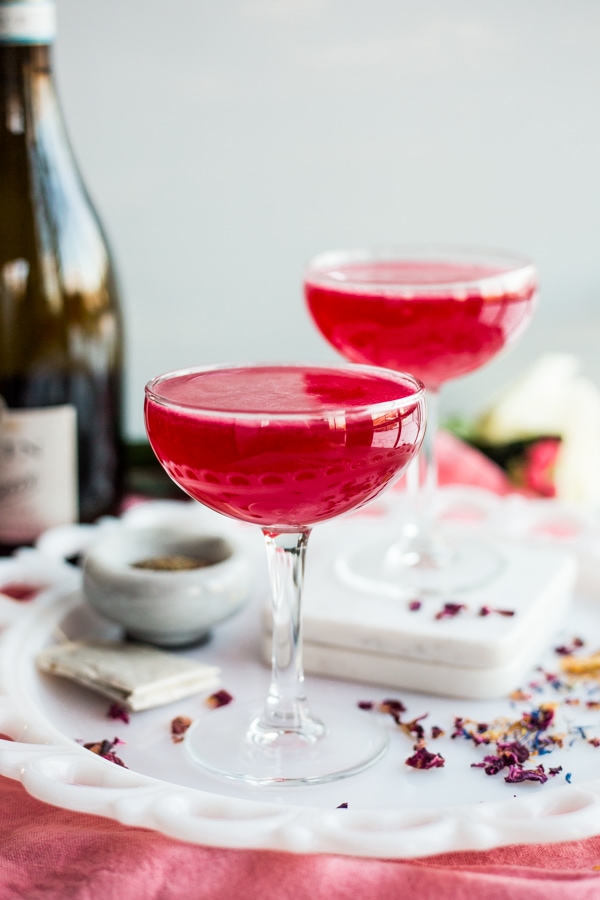 Two coupe glasses filled with a rose hued cocktail with a bottle of Prosecco behind them
