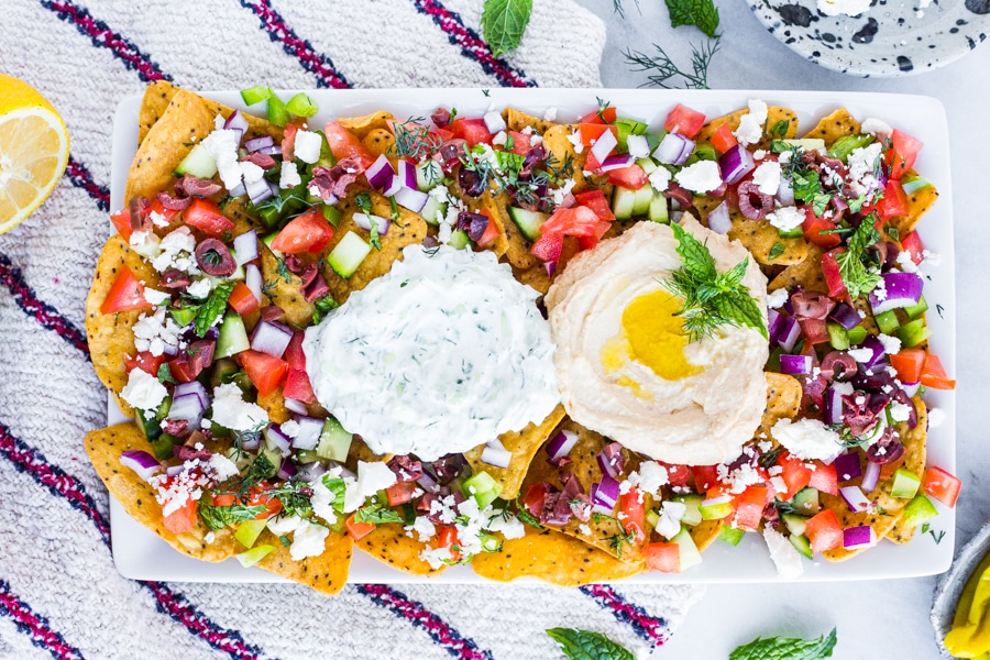 A platter of sweet potato tortilla chips topped with various vegetables, tzatziki, and hummus
