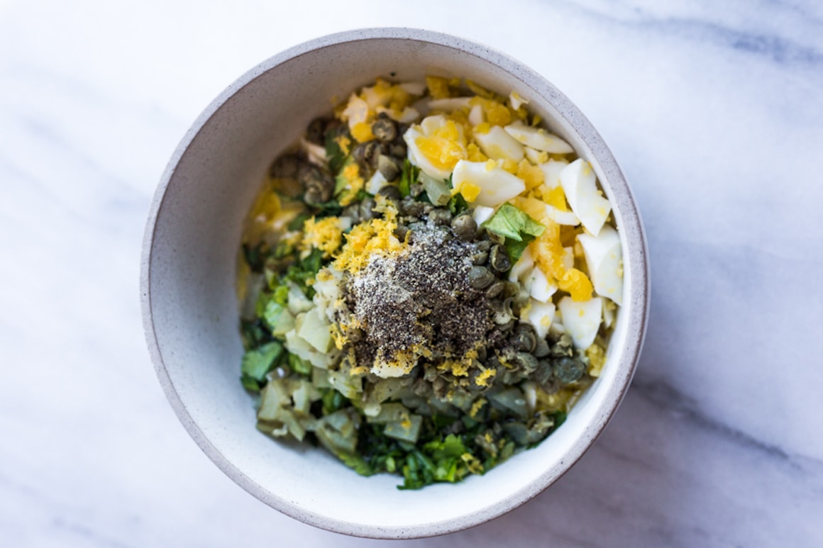 A bowl filled with hardboiled eggs, parsley, chopped cornichons and capers, salt, and pepper
