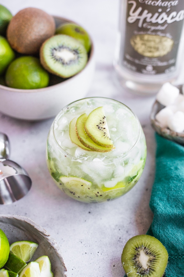 a cocktail topped with kiwi slices surrounded by a bowl of limes and kiwi
