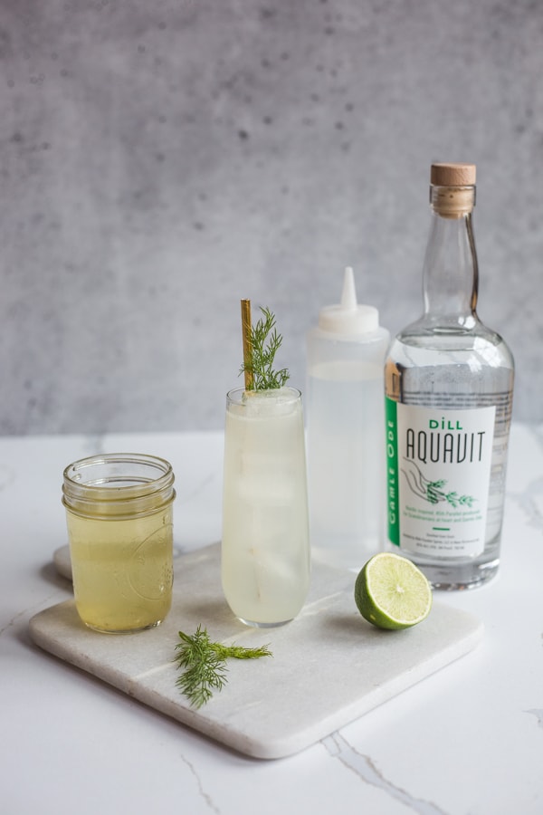 A Tomas Collins cocktail surrounded by a jar of pickle brine, bottle of simple syrup, and bottle of Gamle Ode Dill Aquavit