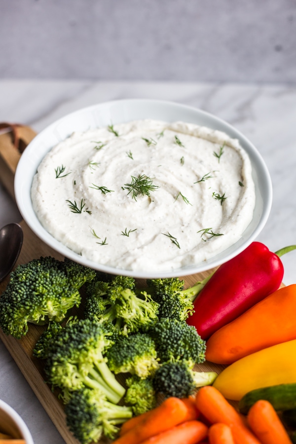 A bowl of ranch dip topped with dill surrounded by vegetables