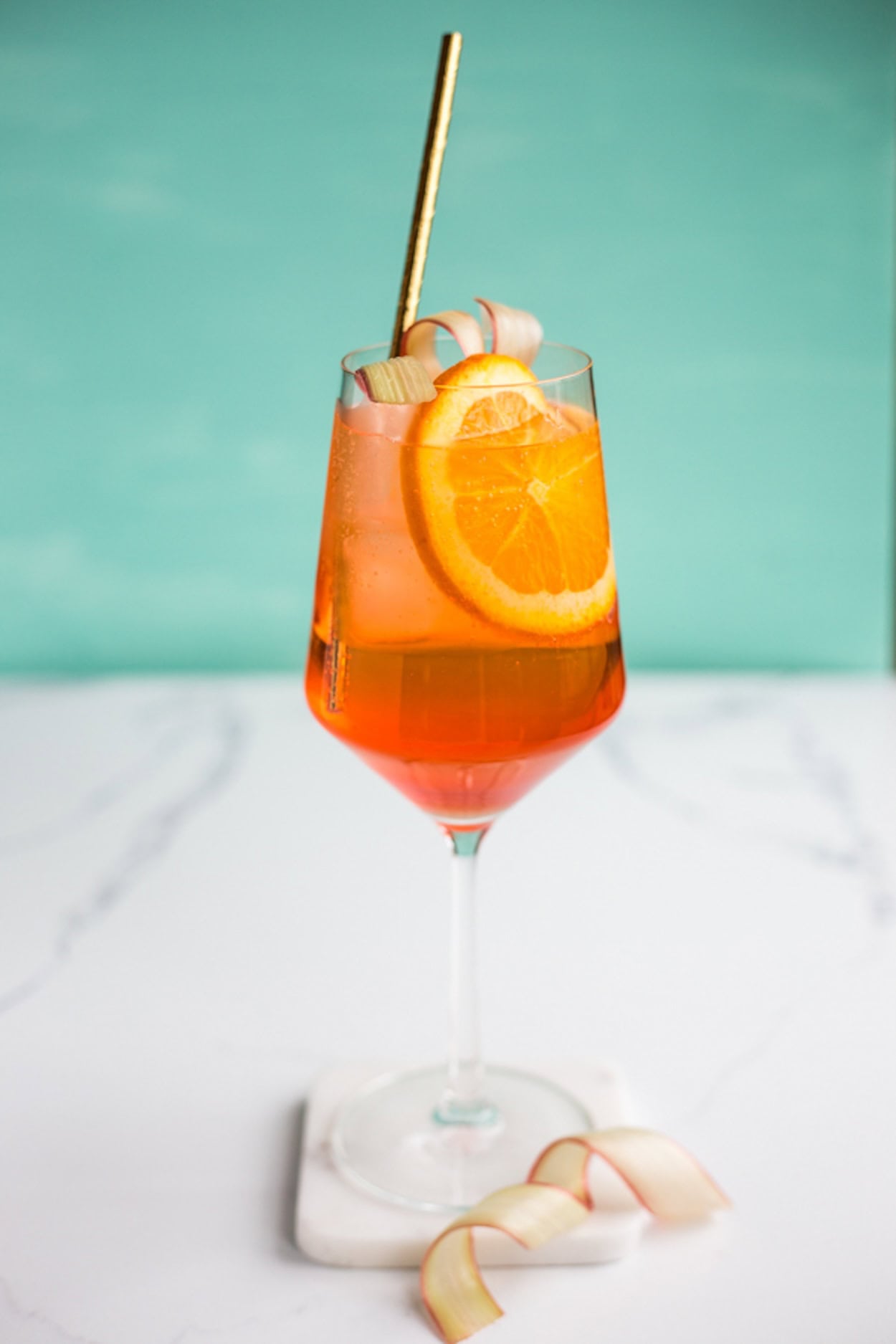 An aperol spritz in a wine glass with a gold straw set against a light blue background