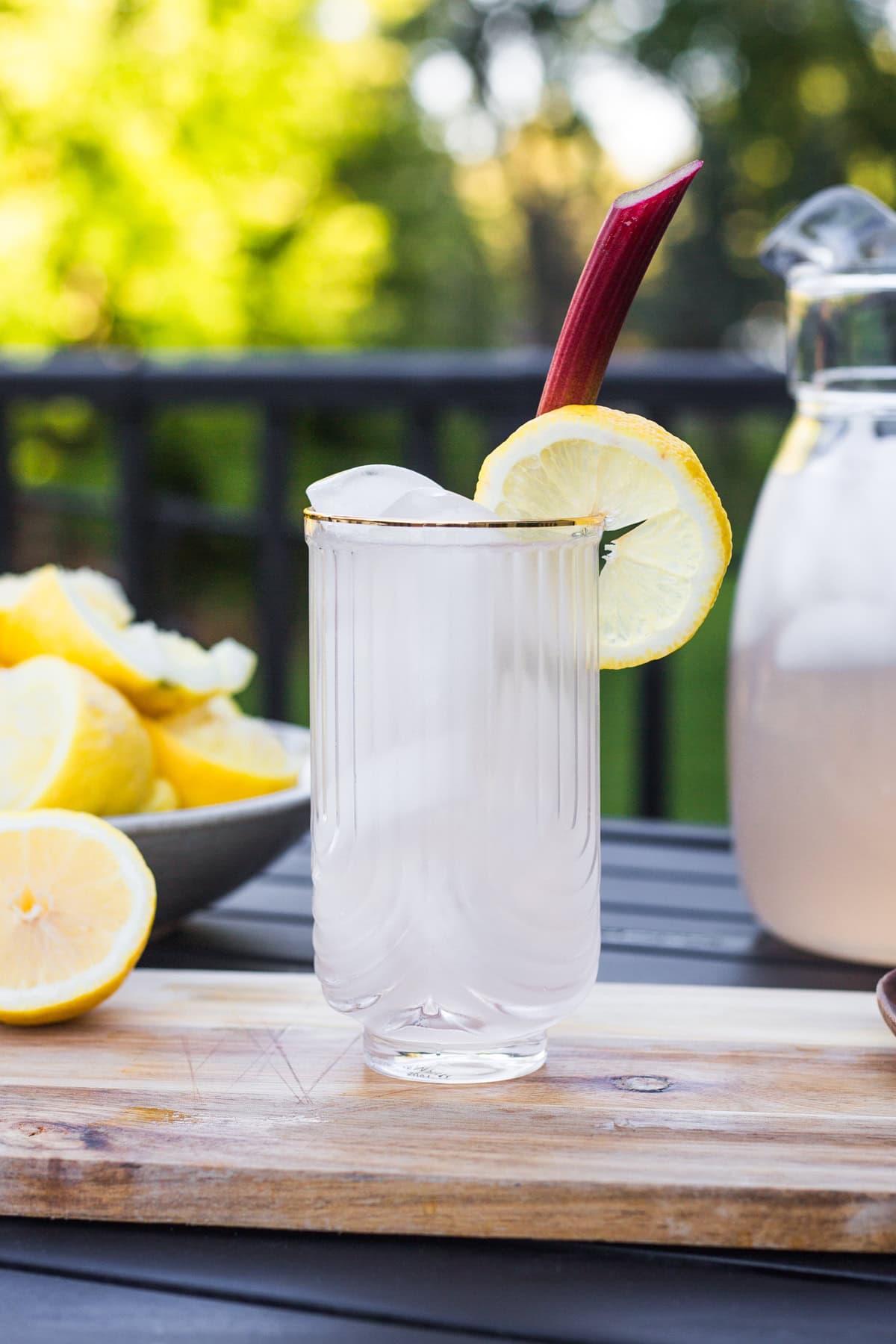 A glass garnished with a rhubarb stalk and lemon wheel with a bowl of lemons and pitcher in the background