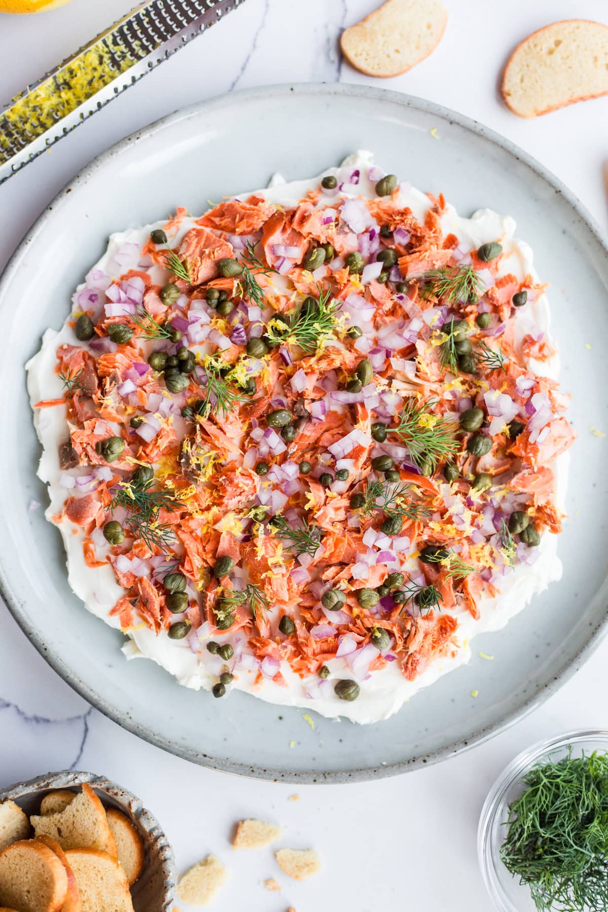 A platter of dip topped with lox, dill, red onion, capers, and dill surrounded by ingredients