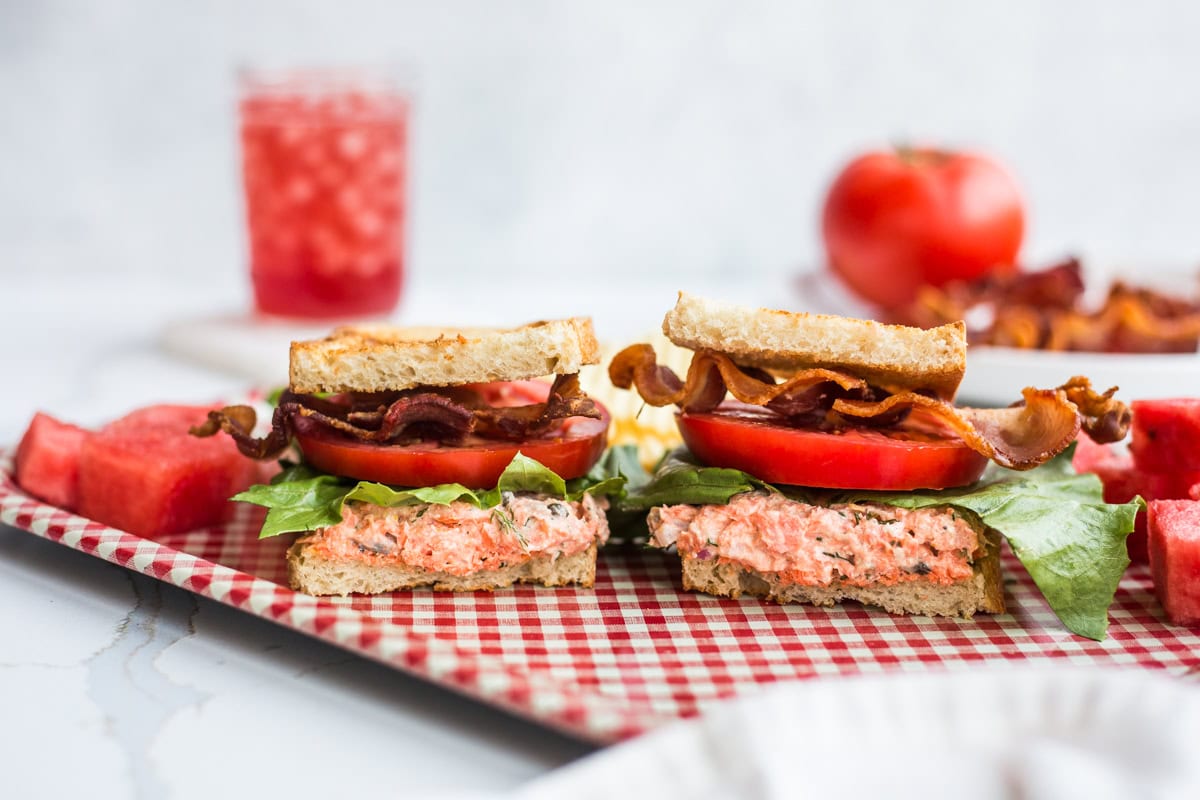 A salmon salad BLT on a red checkered platter with potato chips