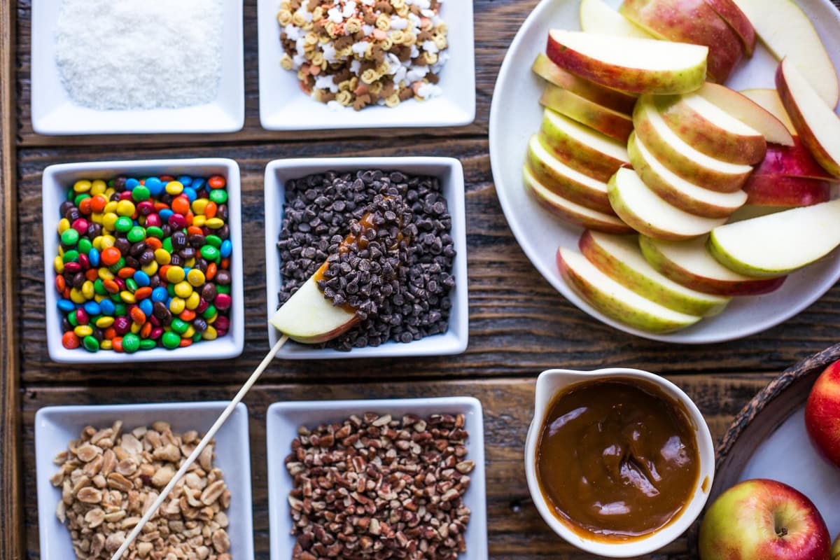 A plate pf apples with a bowl of caramel sauce and a variety of small candies and chopped nuts