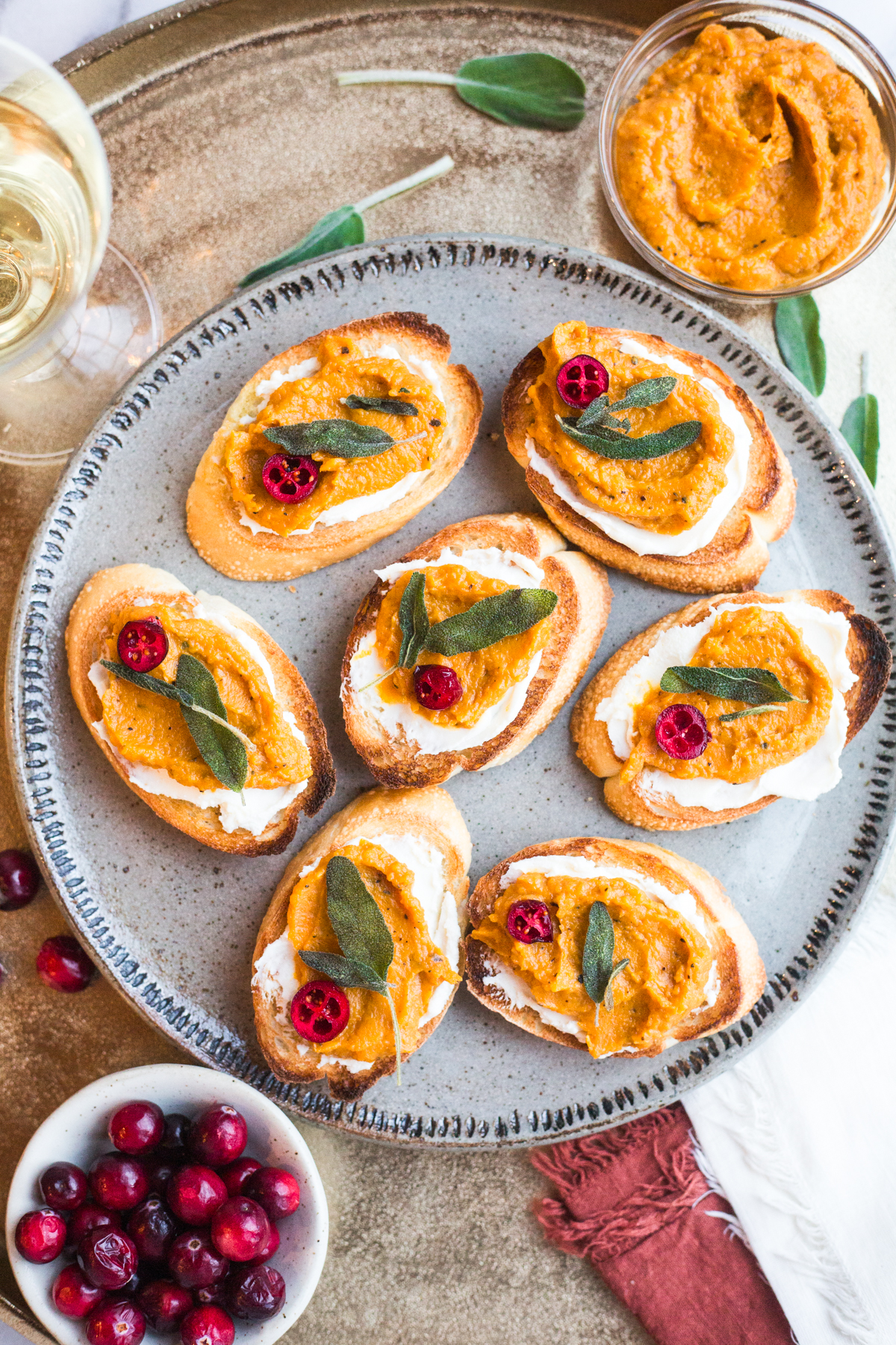 A plate of crostini topped with whipped goat cheese, butternut squash puree, a halved pickled cranberry, and crispy sage