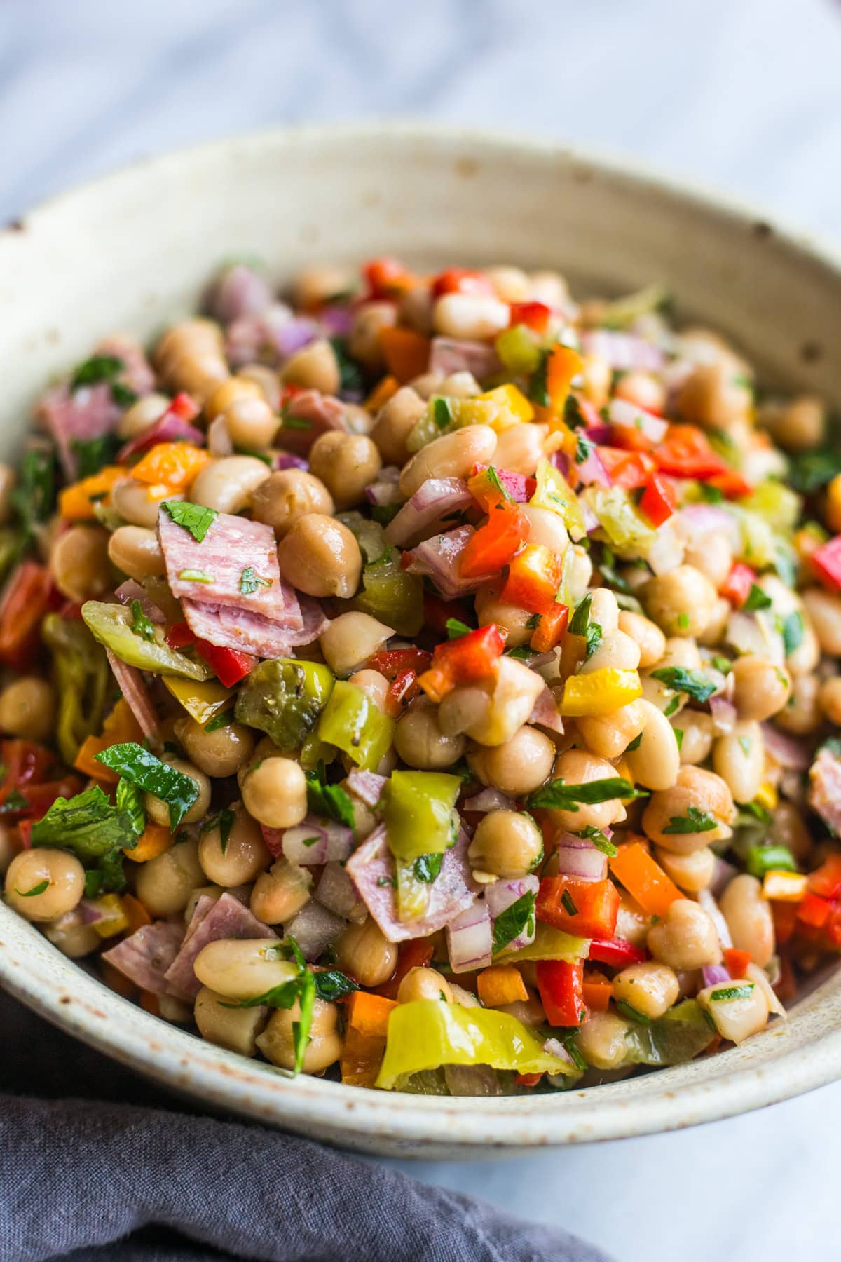 A bowl filled with a bean salad consisting of white beans, garbanzo beans, salami, red onion, peppers, parsley, and pepperoncinis