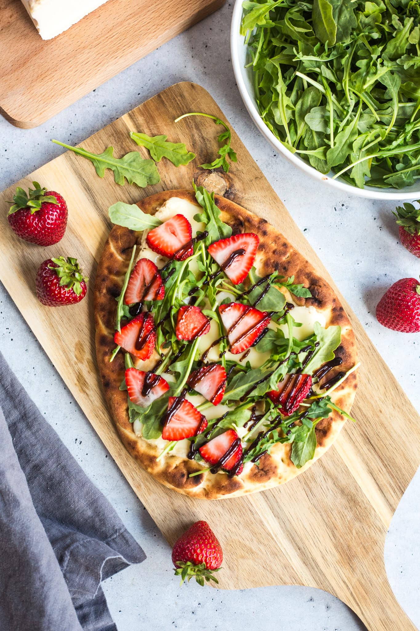 a flatbread topped with cheese, arugula, and strawberries drizzled with balsamic glaze on a wooden platter