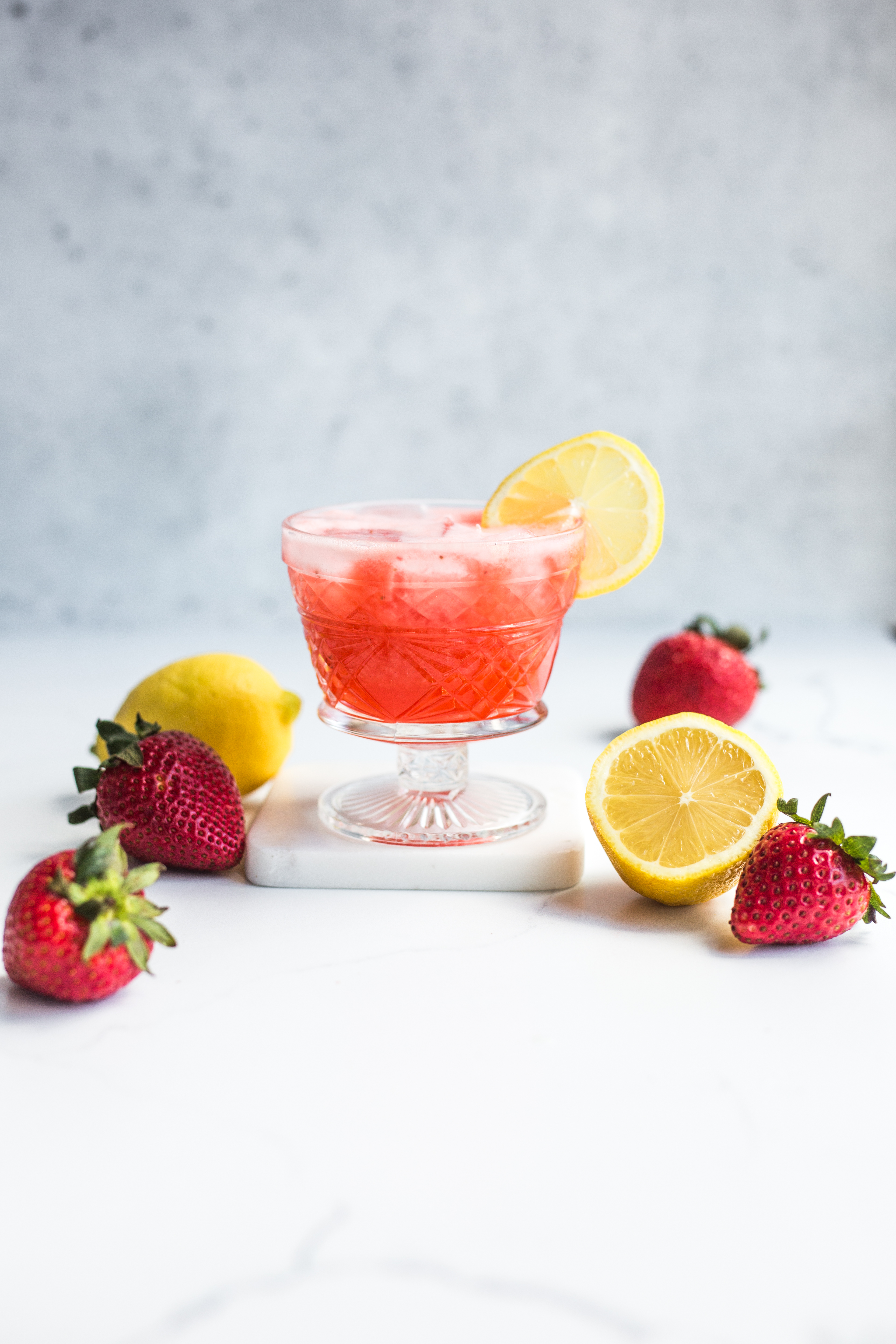 A glass of strawberry lemonade garnished with a lemon slice and surrounded by lemons and strawberries
