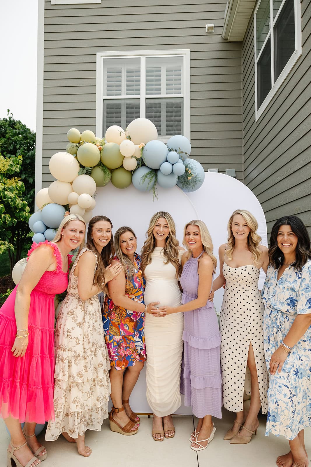 Seven women standing in front of a rounded backdrop outfitted with blue, green, and cream balloons